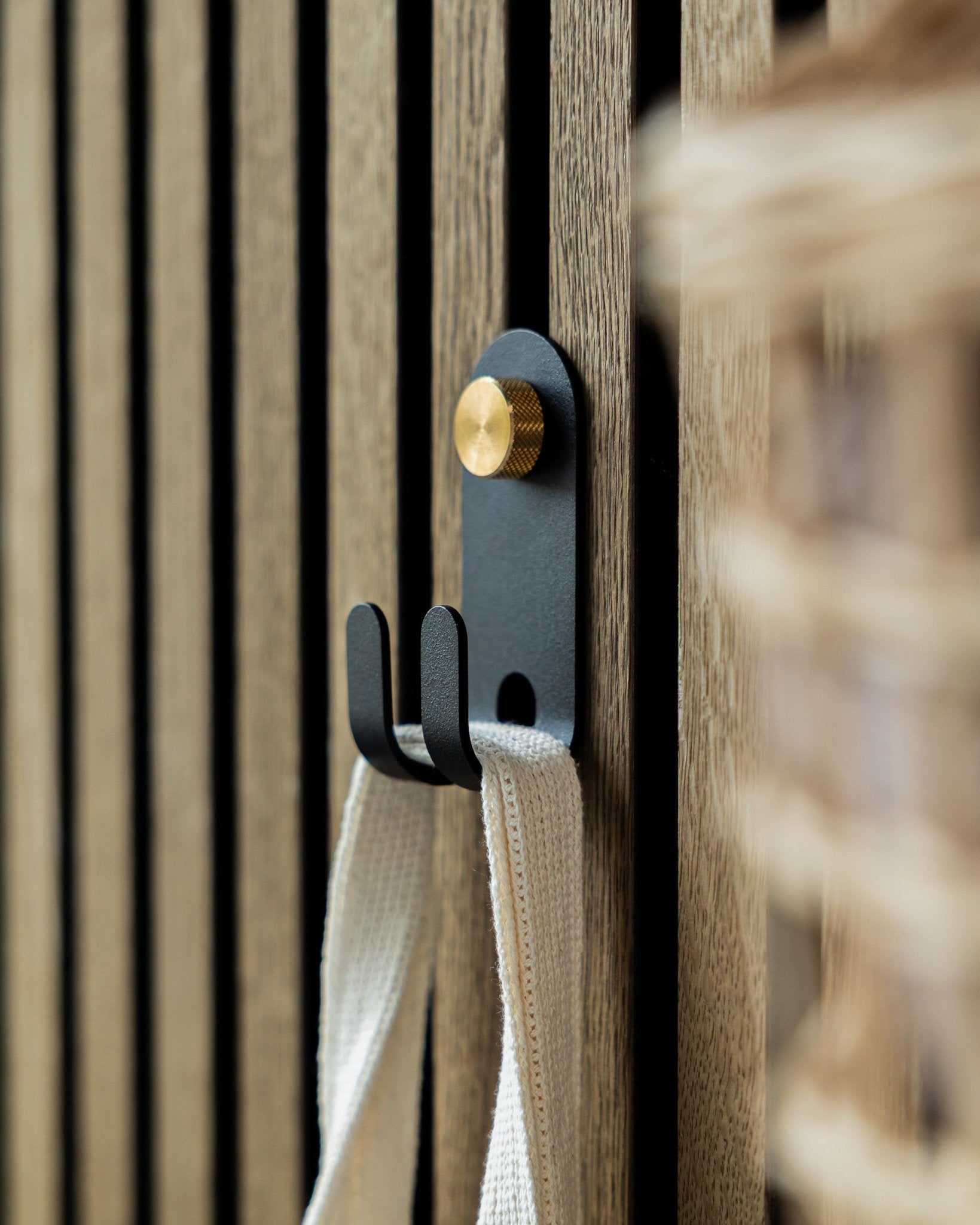 O Norr Double Hanger da WoodUpp em black está montado num painel acústico ripado de madeira Nordic e suporta uma tira em tecido creme-colored.